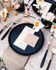 a place setting with black plates, silverware and flowers on the tablecloths