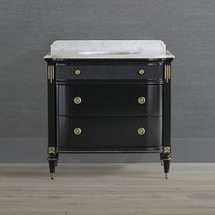 a black dresser with marble top and gold trimmings on the drawers, in front of a gray wall