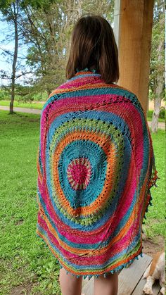 a woman wearing a multicolored crocheted shawl standing in front of a tree