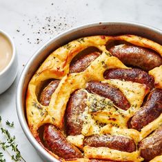 a pan filled with sausages and cheese next to a cup of coffee on a table