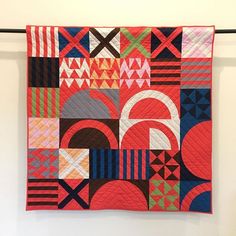 a red and black quilt hanging on a wall in front of a white wall with an abstract design