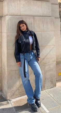 a woman leaning against a wall with her hands on her hips wearing jeans and a leather jacket