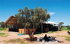 a man sitting under a tree in front of a small building on the side of a dirt road