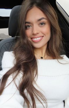 a woman sitting in the back seat of a car with long brown hair and wearing a white sweater