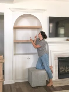 a woman standing on top of a ottoman in front of a tv mounted above a fireplace