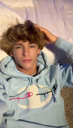 a young man laying on top of a bed wearing a blue sweatshirt and holding his hair in the air