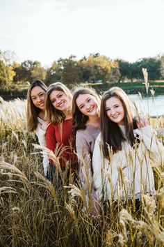 three girls are standing in tall grass by the water and smiling at the camera with their arms around each other