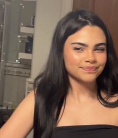 a woman with long black hair standing in front of a kitchen counter and cabinets, smiling at the camera