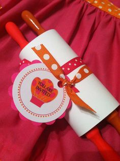 an orange handled spatula with a note attached to it on a pink cloth background