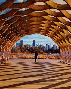 a man walking under a wooden structure in the middle of a park with tall buildings in the background