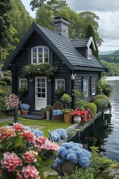 a black house with flowers and potted plants in front of it on the water