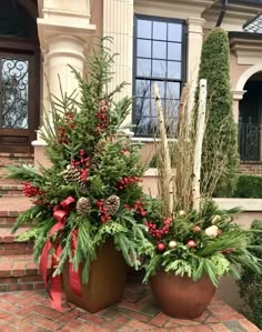 two large planters with evergreens, pine cones and red berries are on the brick walkway in front of a house