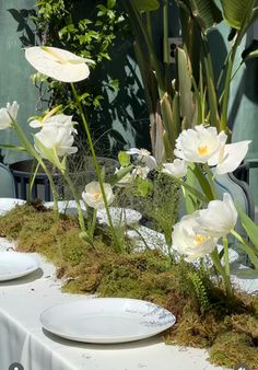 the table is set with white plates and flowers on top of moss covered tablescloths