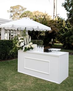 an outdoor bar with white flowers and wine glasses on the counter under an umbrella in front of a house