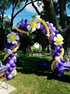 a purple and yellow balloon arch with flowers on it in the middle of a lawn