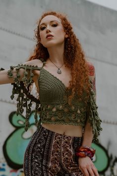 a woman with red hair wearing a green crop top and patterned pants standing in front of a graffiti wall