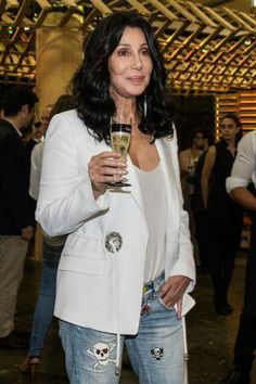 a woman in white jacket and ripped jeans holding a wine glass with people standing around