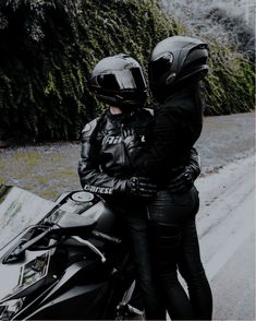 two people on a motorcycle wearing helmets and black leather pants, standing next to each other