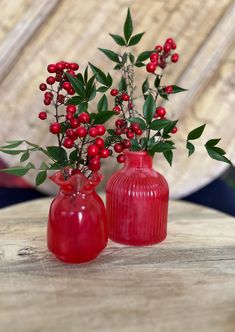 two red vases with berries and greenery in them sitting on a wooden table