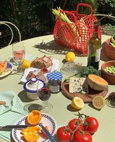 an outdoor table with food and wine on it, including tomatoes, oranges, cucumbers, lemons, cheese, bread