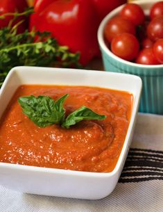 a bowl of tomato sauce next to bowls of tomatoes and basil on a table cloth