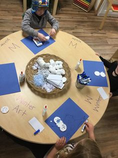 two children are sitting at a table with paper and glue on it, while one child is drawing