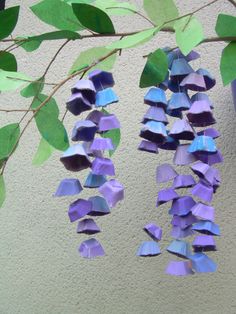 purple and blue paper flowers are hanging from a branch with green leaves on the wall