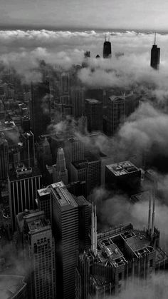 black and white photograph of city buildings in the sky with clouds surrounding them from above