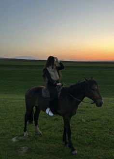 a woman riding on the back of a brown horse in a green field at sunset