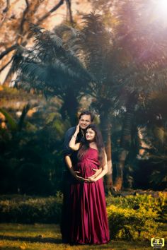 a pregnant woman in a red dress poses for a photo with her husband while the sun shines through the trees