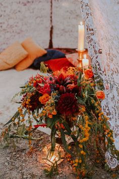 an arrangement of flowers and candles is on the ground next to a wall with lace