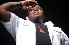 a man in white shirt and necklace holding his hand up to his head while standing on stage