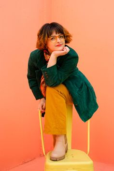 a woman sitting on top of a yellow chair with her hand under her chin and looking at the camera