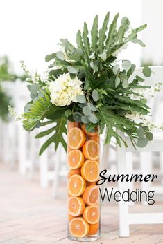 a vase filled with oranges and greenery on top of a brick floor next to a white chair