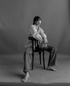 a woman sitting on top of a wooden chair in front of a gray background with her legs crossed