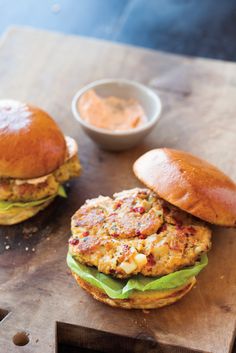 two burgers sitting on top of a wooden cutting board next to a bowl of sauce