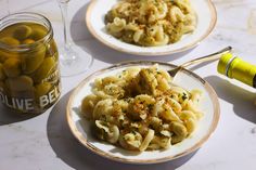 two plates filled with pasta and olives next to a jar of olive oil on a table