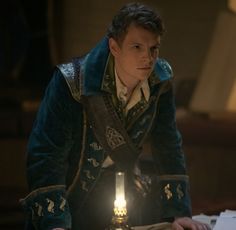 a young man dressed in medieval clothing is looking at something on a table with a lit candle