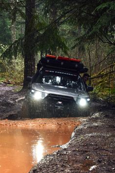 a car driving through a puddle in the woods with its lights on and it's hood up