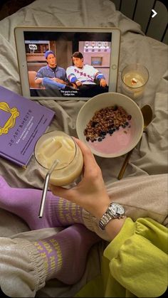 a person laying on a bed with food and a book in front of them while watching tv