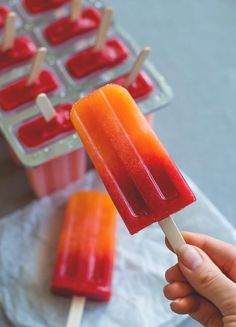 a hand holding an orange and red popsicle next to trays of ice cream