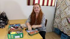 a woman sitting at a table with many items in her hand and an open suitcase on the floor