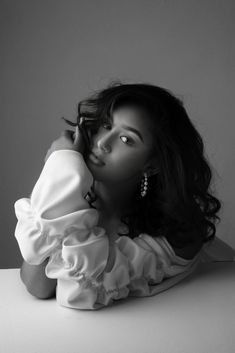 a woman sitting on top of a table next to a white wall with her hand under her chin