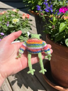 a hand holding a small knitted frog in front of potted plants and flowers