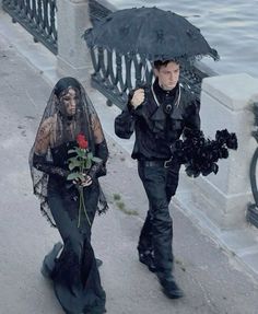 two people dressed in black walking down the street with an umbrella over their heads and one holding a rose