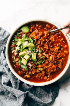 a person holding a spoon full of chili and avocado in a white bowl