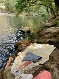 there is a blanket, book and shoes on the rocks by the riverbank with trees in the background