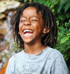 a young boy with dreadlocks laughing in front of a waterfall and greenery