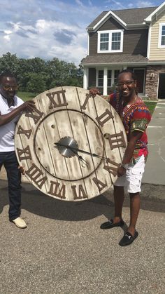 two men standing next to each other holding a large clock in front of a house