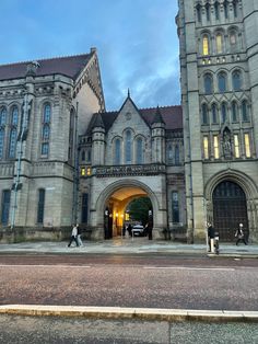 two people walking in front of an old building at dusk with cars parked outside the entrance